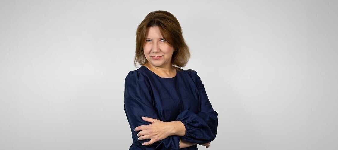 a woman in a dark dress with her arms folded, against a light, plain background