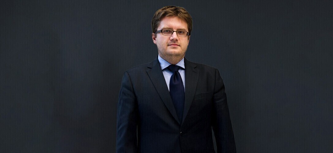 Męzczyzna w białej koszuli, ciemnym krawacie i ciemnej marynarce na jednolitym ciemnym tle