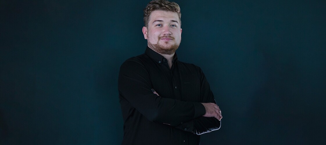 Mężczyzna w ciemnej koszuli, stoi z założonymi rękami na ciemnym jednolitym tle