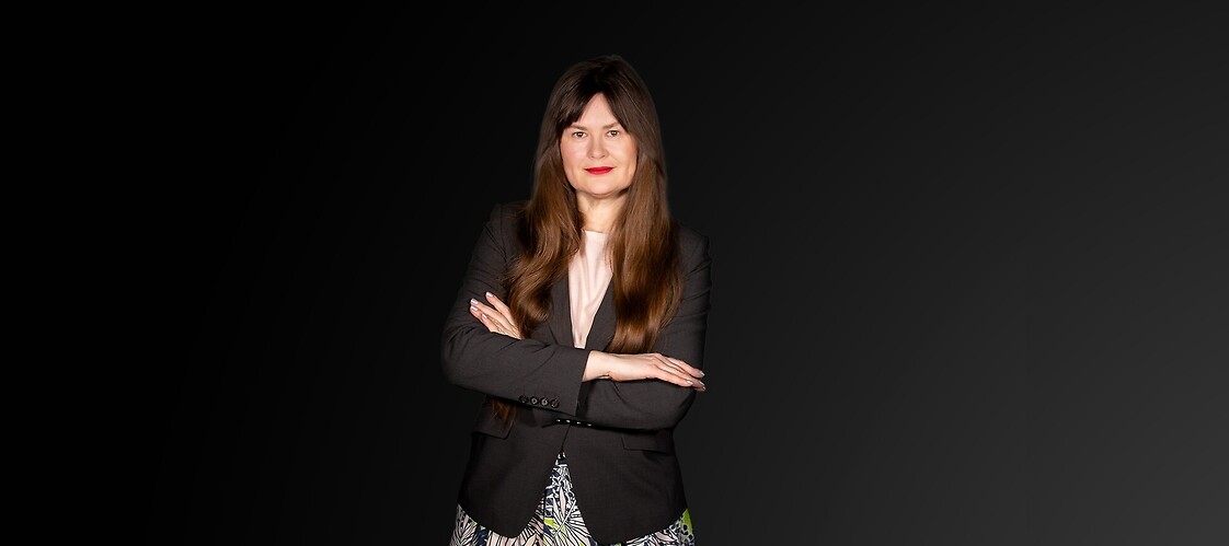 Kobieta w jasnej bluzce, ciemnej marynarce i wzorzystej spódnicy na ciemnym jednolitym tle