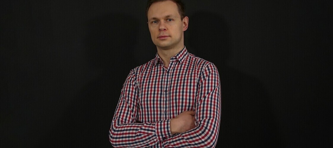 Mężczyzna w koszuli w kratkę, z założonymi rękami na ciemnym, jednolitym tle