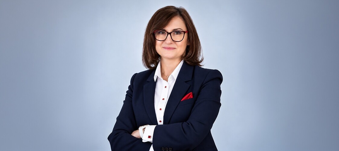 LPbieta w ciemnym żakiecie i jasne bluzce, z założonymi rękami, na jasnym tle