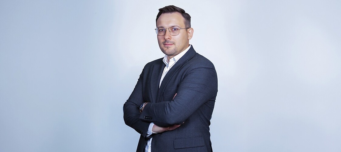 Mężczyzna w  marynarce i jasnej kouli, z założonymi rękami, na jasnym tle