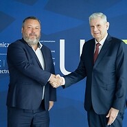 Dwóch mężczyzn w garniturach wita sie uściskiem dłoni na tle z napisem ...