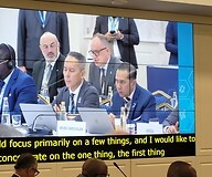 Kadr z telebimu: grupa mężczyzn za stołami konferencyjnymi, w drugim rzędzie ...