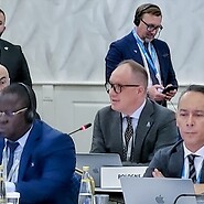 Grupa mężczyzn za stołami konferencyjnymi, w drugim rzędzie przemawiający mężczyzna