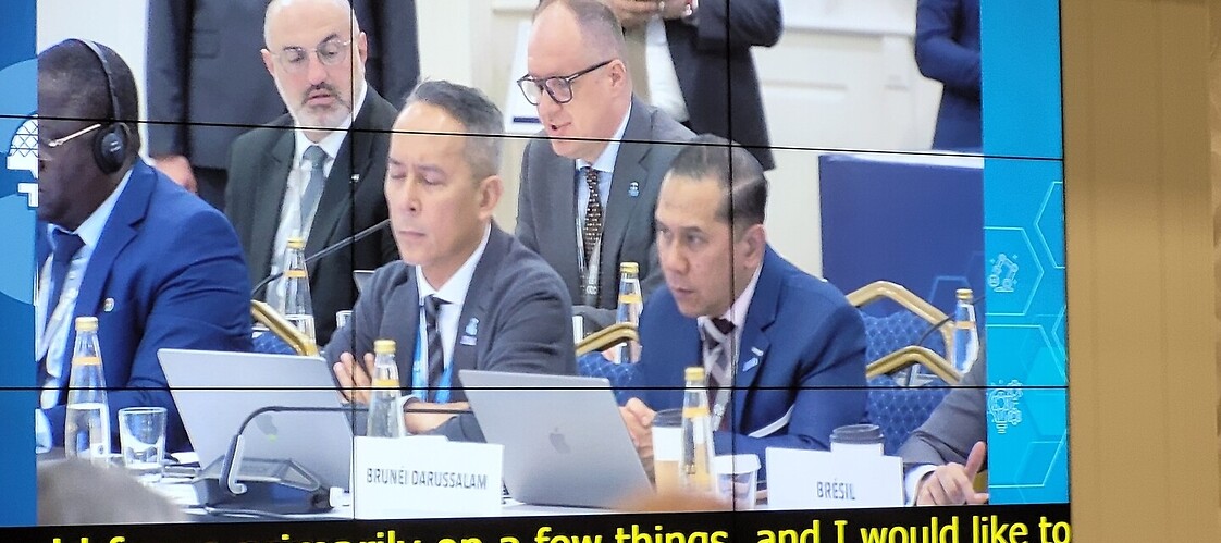 Kadr z telebimu: grupa mężczyzn za stołami konferencyjnymi, w drugim rzędzie przemawiający mężczyzna