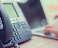 Telefon stacjonarny, w tle dłonie na klawiaturze kmpuera
