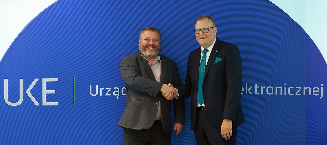 Dwóch mężczyzn w garniturach podaje sobie dłoń na tle bannera z logotypem UKE