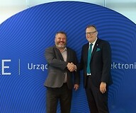 Dwóch mężczyzn w garniturach podaje sobie dłoń na tle bannera z logotypem UKE