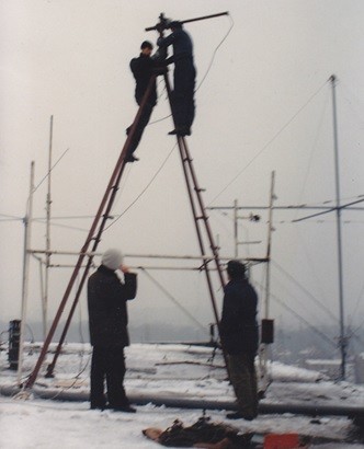 Montaż anteny logarytmiczno-periodycznej - dwóch pracowników na drabinie montuje urządzenia. Na dole dwóch pracowników nadzorujących pracę.